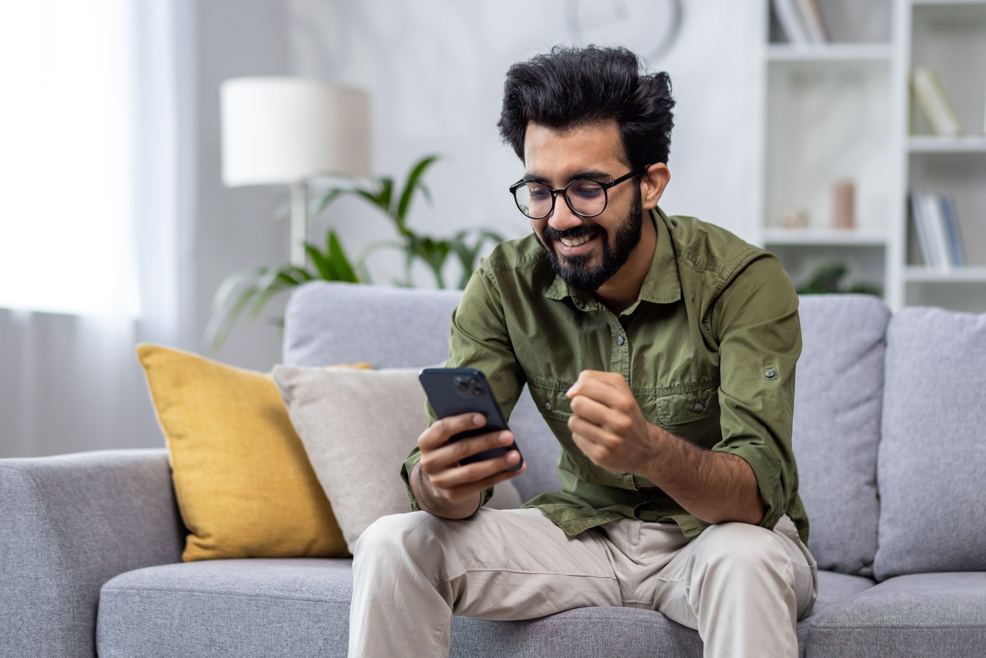 Happy and satisfied with achievement results hispanic at home reading online win notification in online application on phone, man smiling holding hand up triumph gesture sitting sofa in living room